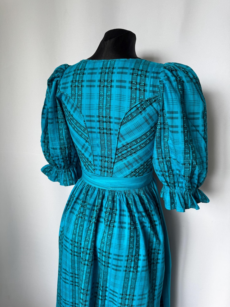 Puede incluir: Vestido vintage de color turquesa con estampado de cuadros, con mangas abullonadas y corpi&ntilde;o entallado. El vestido tiene una faja ancha turquesa en la cintura y pu&ntilde;os con volantes. El vestido se exhibe en un maniqu&iacute; negro.