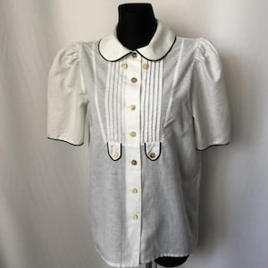 May include: A white button-down shirt with a black trim collar and cuffs. The shirt has a pleated front and a single button closure.