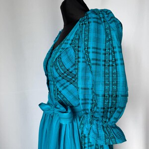 Puede incluir: Un vestido turquesa con un estampado de cuadros negros y turquesas. El vestido presenta un cuello en V, mangas abullonadas y un lazo en la cintura. La falda tiene rayas verticales. El vestido se exhibe en una cabeza de maniqu&iacute; negra.