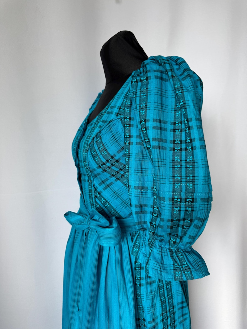 Puede incluir: Un vestido turquesa con un estampado de cuadros negros y turquesas. El vestido presenta un cuello en V, mangas abullonadas y un lazo en la cintura. La falda tiene rayas verticales. El vestido se exhibe en una cabeza de maniqu&iacute; negra.