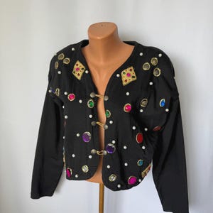 Op de afbeelding: Zwarte jas met lange mouwen, versierd met kleurrijke edelstenen, gouden versieringen en witte parelaccenten. De jas heeft een haak-en-oogsluiting en wordt gepresenteerd op een mannequin. Een uniek, opvallend kledingstuk.