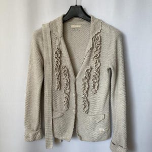 May include: A beige knitted cardigan with a ruffled trim down the front and a matching belt. The cardigan has a button closure and a pocket on the left side.