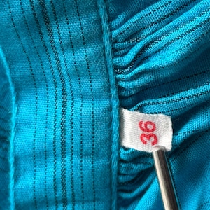 Puede incluir: Primer plano de una tela turquesa con rayas negras verticales. Una etiqueta blanca con el n&uacute;mero "36" en rojo est&aacute; adjunta. La tela est&aacute; fruncida, creando una apariencia texturizada.