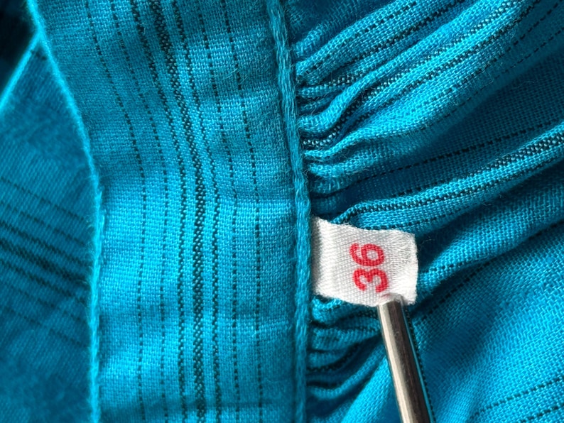Puede incluir: Primer plano de una tela turquesa con rayas negras verticales. Una etiqueta blanca con el n&uacute;mero "36" en rojo est&aacute; adjunta. La tela est&aacute; fruncida, creando una apariencia texturizada.