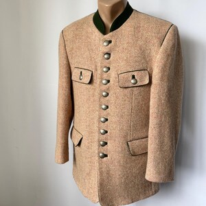 May include: A tan herringbone blazer with a dark green collar and black trim. The blazer has silver buttons down the front and on the pockets. The jacket is displayed on a mannequin.