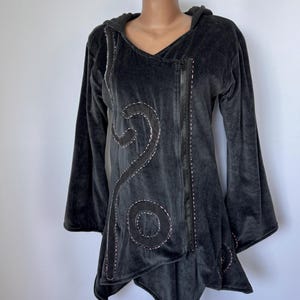 Könnte beinhalten: Eine dunkelgraue Kapuzenjacke aus Velours mit asymmetrischem Frontreißverschluss. Das Kleidungsstück ist mit schwarzen Stoffapplikationen in wirbelnden und kreisförmigen Formen verziert, die durch sichtbare weiße Steppnähte betont werden.