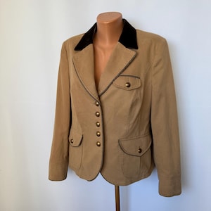 Puede incluir: Un blazer color beige con cuello de terciopelo negro, cuatro bolsillos delanteros y botones dorados. El blazer tiene ribetes marrones oscuros y se exhibe en un maniquí. Una chaqueta clásica y entallada.