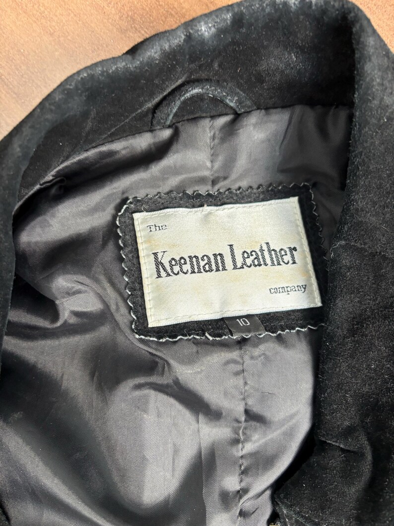 May include: Close-up of a black leather jacket's interior. The lining is black, and a label reads "The Keenan Leather company." The label is white with black text and a decorative border. A size 10 tag is visible.