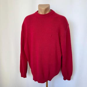 May include: A solid red long-sleeve jumper featuring a classic crew neckline, ribbed cuffs, and a ribbed hem. The knit garment has a simple, clean design with a consistent deep red colour throughout the entire piece, including the collar.