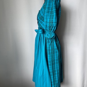 Puede incluir: Un vestido turquesa con un estampado de cuadros en el corpi&ntilde;o y las mangas, y una falda turquesa lisa. El vestido tiene una cintura entallada con un detalle de lazo, y las mangas est&aacute;n recogidas en los pu&ntilde;os. El vestido se exhibe en un maniqu&iacute;.