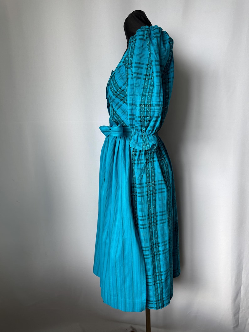 Puede incluir: Un vestido turquesa con un estampado de cuadros en el corpi&ntilde;o y las mangas, y una falda turquesa lisa. El vestido tiene una cintura entallada con un detalle de lazo, y las mangas est&aacute;n recogidas en los pu&ntilde;os. El vestido se exhibe en un maniqu&iacute;.