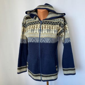 May include: A long-sleeve zip-up hooded jacket featuring a dark blue lower section and a cream-coloured upper section with a repeating pattern of llamas and geometric shapes in shades of brown, black, and blue. The jacket has a full-length front zip.