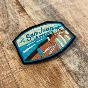 Peut inclure: Écusson brodé représentant une scène côtière avec le texte "San Juan La Union". L'écusson présente une palette de couleurs bleu, marron et beige, avec un palmier et des planches de surf. L'écusson est bordé de noir et est posé sur une surface en bois.