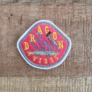 Può includere: Toppa ricamata a forma di diamante rosso, con le parole "DRAGON VIBES" in giallo, con un design stilizzato di drago in blu e viola. La toppa ha un bordo bianco ed è posta su uno sfondo di legno.