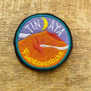 Peut inclure: Un patch rond brodé avec un croissant de lune et les mots "TINAYA Fuerteventura" en jaune. Le fond est violet avec une chaîne de montagnes en orange et rouge.