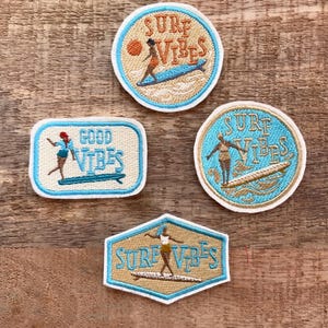 Può includere: Quattro toppe ricamate con un design di una ragazza che fa surf. Le toppe sono blu, beige e dorate con il testo "Surf Vibes" e "Good Vibes".