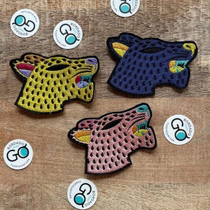 Può includere: Tre toppe ricamate con teste di animali colorate con un motivo ripetitivo. Ogni toppa ha un adesivo circolare con il testo "BORDADO GO FUERTEVENTURA" in blu e bianco.