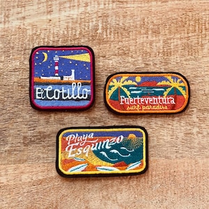 Può includere: Tre toppe ricamate con disegni e testo colorati. La prima toppa è blu con un faro e il testo "El Cotillo". La seconda toppa è rossa con palme e il testo "Fuerteventura surf paradise". La terza toppa è arancione con un tramonto e il testo "Playa Esquinzo".