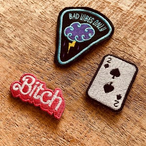 Può includere: Tre toppe ricamate su una superficie di legno. Una toppa recita "Bad Vibes Only" con un design di nuvola e fulmine. Un'altra toppa dice "Bitch" in corsivo rosso. La terza toppa è una carta da gioco, il due di picche.