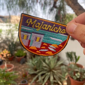 Può includere: Una toppa ricamata colorata con il testo "Majanicho" in lettere gialle. La toppa presenta un'immagine stilizzata di una casa con un tetto turchese e accenti arancioni.