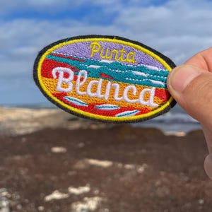 Può includere: Una toppa ricamata colorata con il testo "Punta Blanca" in un design giallo, rosso e blu. La toppa è di forma ovale e ha un bordo nero.
