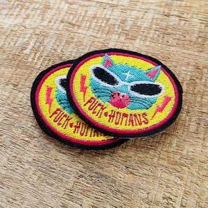 May include: Two round yellow embroidered patches with black outlines. The patches feature a blue cat wearing sunglasses and the text "F*CK HUMANS" in pink.
