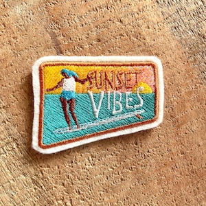 Peut inclure: Un patch brodé avec une silhouette de surfeur sur une planche de surf, un coucher de soleil en arrière-plan et le texte "SUNSET VIBES".