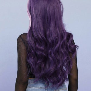May include: A long, wavy purple wig, approximately 28 inches in length, is shown from the back. The wig has a deep purple hue with a slight pinkish undertone. The model is wearing a black mesh top and blue jeans.