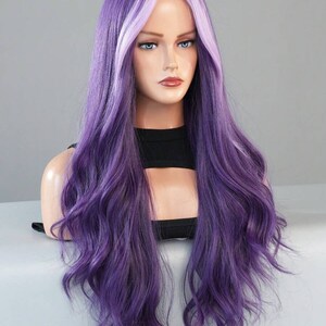 May include: A long, wavy wig in shades of purple and lavender is displayed on a mannequin head. The wig features a dark purple base with lighter lavender highlights framing the face. The hair is styled with loose waves, creating a voluminous and natural look.