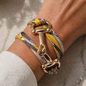 Könnte beinhalten: Armband aus goldfarbenem Metall mit einem Gebiss-Design. Das Armbandband besteht aus einem bunten Stoff mit gelben, rosa und grauen Mustern. Das Armband ist um ein Handgelenk gewickelt.