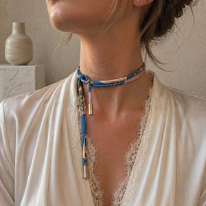 Könnte beinhalten: Ein blau-rosa Seidenschal mit Blumenmuster, der um den Hals gebunden ist. Der Schal hat goldfarbene Metallakzente an den Enden. Die Person trägt einen weißen Satin-Bademantel mit Spitzenbesatz.