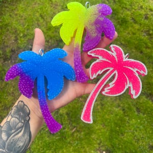 May include: Three palm tree ornaments made of resin. One is blue and purple, one is neon yellow and purple, and one is bright pink with silver glitter edges. Each ornament features a small metal loop at the top for hanging purposes.