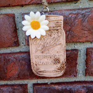 May include: A rustic, distressed, light brown mason jar ornament with a white daisy attached. The jar has the text "Ball Perfect Mason" printed on it.