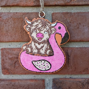 May include: A pink glitter flamingo pool float with a brown and white highland cow sitting inside. The float has black outlines and pink and white polka dots.