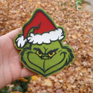 Puede incluir: Primer plano de una cara verde del Grinch con un gorro de Papá Noel rojo y ribete blanco. El Grinch tiene ojos amarillos y un contorno negro. Decoración navideña.
