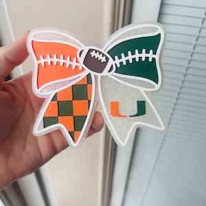 Puede incluir: Un lazo decorativo en colores naranja, verde y marrón. El lazo tiene un balón de fútbol en el centro y la letra "U" en un lado. El lazo está sostenido por una mano.