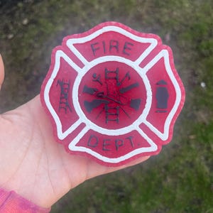 Op de afbeelding: Een rood en wit brandweerembleem met de tekst "FIRE DEPT.". Het embleem heeft een kruis van Malta-ontwerp met zwarte gereedschappen in het midden. Het embleem wordt in een hand gehouden.