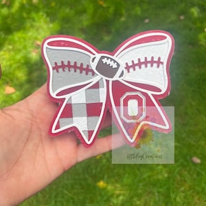 May include: A layered bow-shaped ornament featuring a red, white, and gray color scheme. The design includes a football graphic, a checkered pattern, and a letter O emblem. The edges are outlined in red, and the surface has a textured appearance.