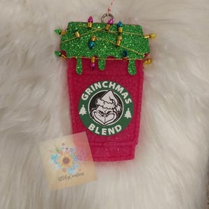May include: A pink and green glitter Christmas ornament shaped like a coffee cup with the text "Grinchmas Blend" and a green Grinch face logo.