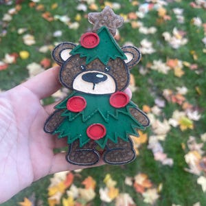 Op de afbeelding: Een kerstboomvormige berenornament. De bruine beer heeft een groen boomvormig lichaam met rode cirkelvormige ornamenten. Een ster bekroont de boom. Het ornament wordt tegen een achtergrond van groen gras en gevallen bladeren gehouden.