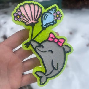 Könnte beinhalten: Ein neongrüner Lufterfrischer in Form eines grauen Delfins mit einer rosa Schleife, der rosa und blaue Luftballons hält. Die Ballons sind mit Blumen gekrönt. Eine rosa Muschel ist ebenfalls befestigt.