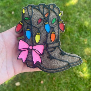 Può includere: Un ornamento a forma di stivale da cowboy marrone glitterato con un contorno nero. Lo stivale è decorato con luci natalizie colorate e un fiocco rosa acceso. L'ornamento è tenuto in mano su uno sfondo verde.