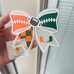 May include: A decorative bow-shaped ornament with orange, green, and brown accents. The bow features a football in the centre and the letters "U" and "L". The bow is held by a hand, and a small sticker with text is visible.