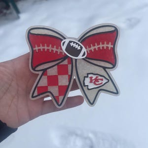 May include: A decorative bow with a football theme. The bow features a red and white checkered pattern, a red section with white stitching, and a football in the centre. The bow also has the Kansas City Chiefs logo.