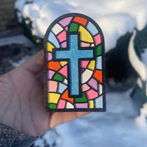 May include: A colorful, stained-glass-style cross with a blue cross at the center. The cross is surrounded by a mosaic of vibrant colors including pink, orange, yellow, and green. The frame is black.