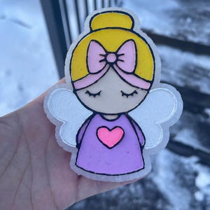 May include: A decorative angel figure with a yellow bun and a pink bow. The angel has closed eyes, a purple dress with a pink heart, and white wings. The outline is black.