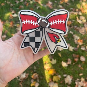 May include: A decorative bow with a football and team emblem design. The bow features a red top section with white stitching, a brown football, a black and white chequered section, and a red bird logo.