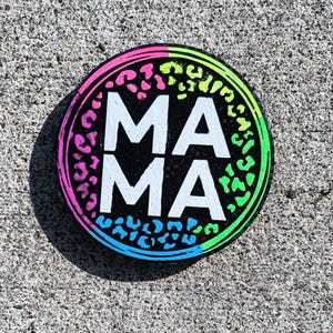 May include: A round black and white sign with the word "MAMA" in white letters. The sign has a pink, green, and blue leopard print border.