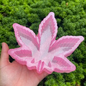 May include: A pink, glittery cannabis leaf-shaped tray. The tray has a layered design with a white center and a pink, glittery border. The tray is held in a hand against a backdrop of green foliage.