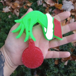 Puede incluir: Una mano verde del Grinch sosteniendo un adorno navideño rojo. El Grinch lleva un gorro de Papá Noel rojo y blanco.